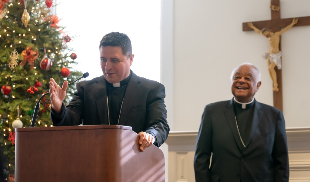 A Latino bishop speaks from behind a lectern. A Christmas tree, a crucifix, and a beaming Cardinal Wilton Gregory, an African-American man, stand behind him.