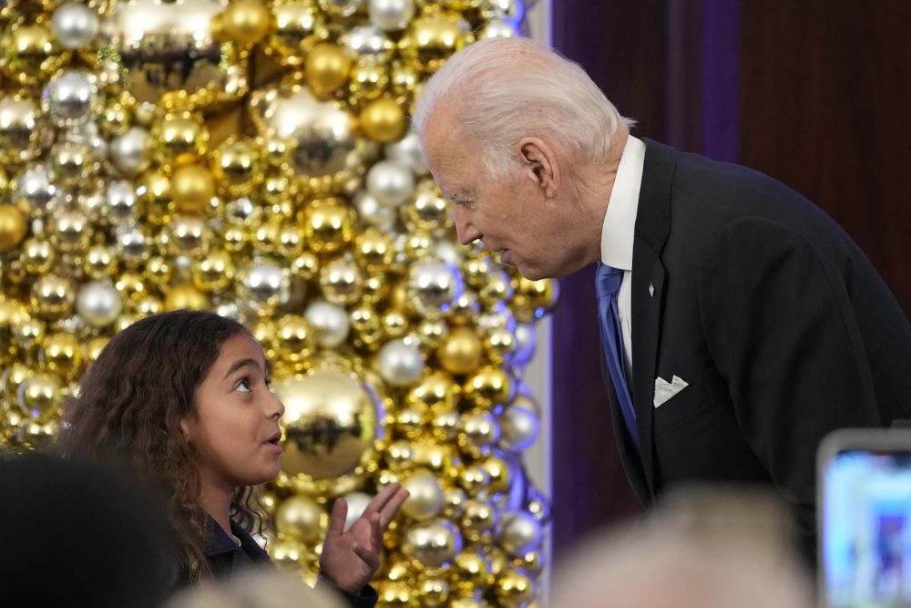 Biden leans down towards a girl with brown curly hair. They stand in front of silver and gold orbs