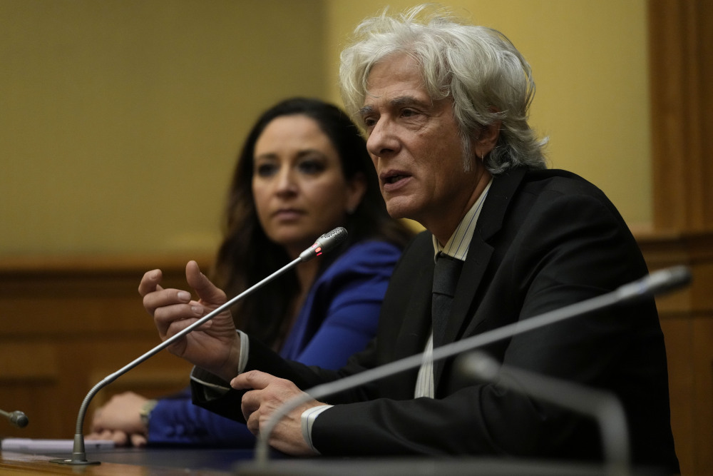 A man with floppy white hair sits next to a woman with brown hair behind microphones