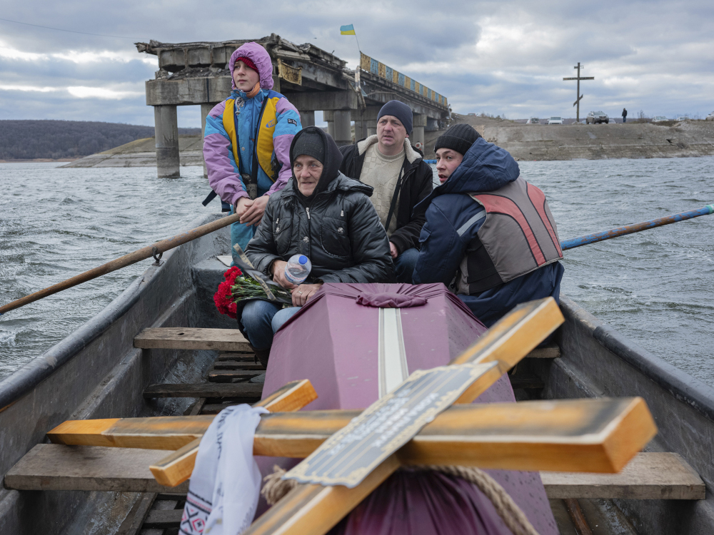 Four people with winter coats sit in a rowboat with a coffin and large cross