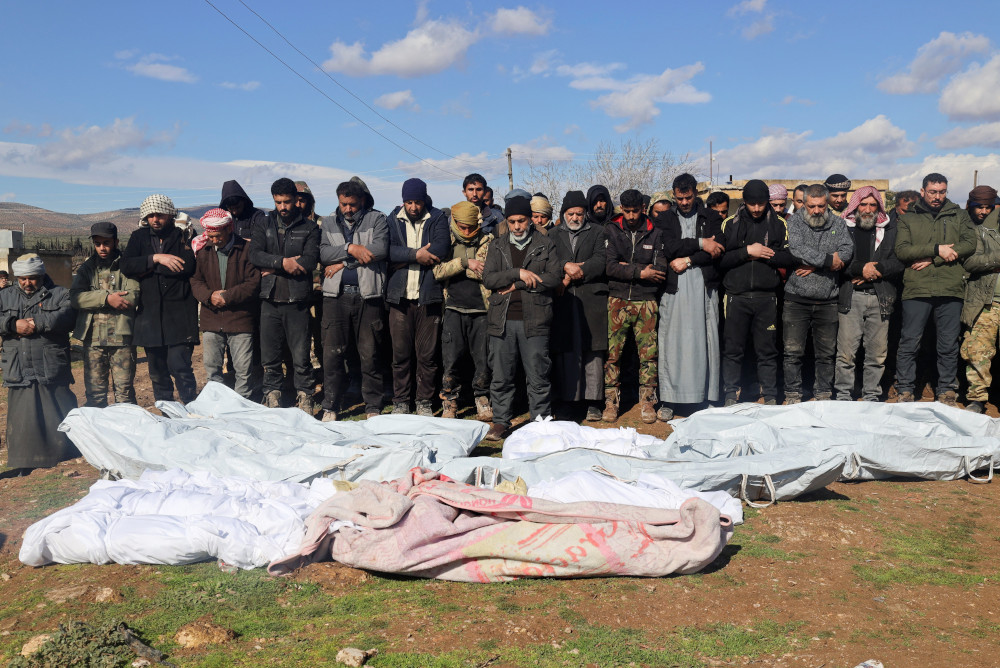 A group of people stand outside around bodies fully wrapped in cloth