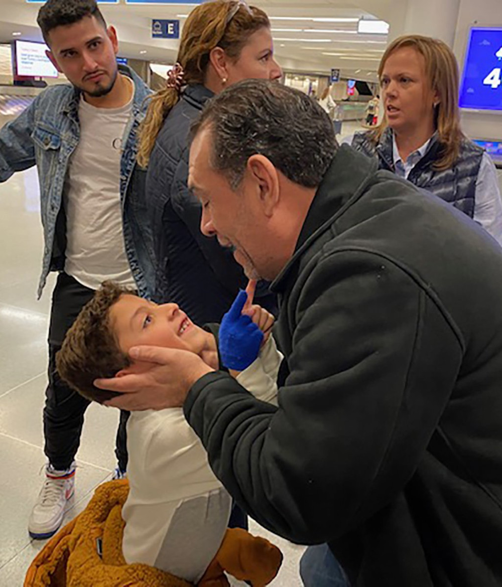 A man in a black jacket grips a little boy's face with both hands as the little boy beams up at him