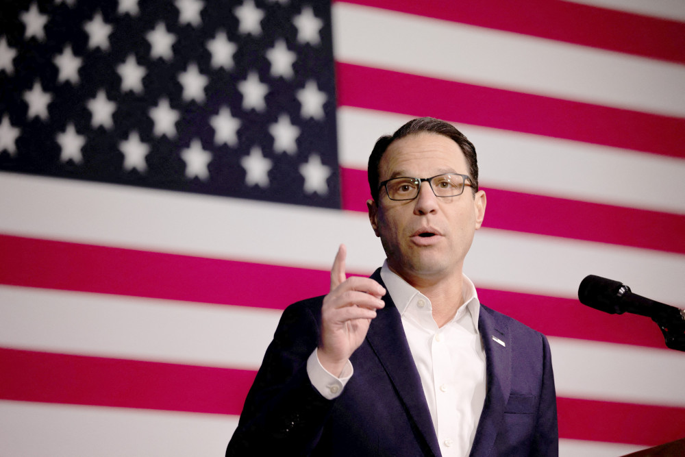 A white man with glasses stands behind a microphone and in front of an American flag