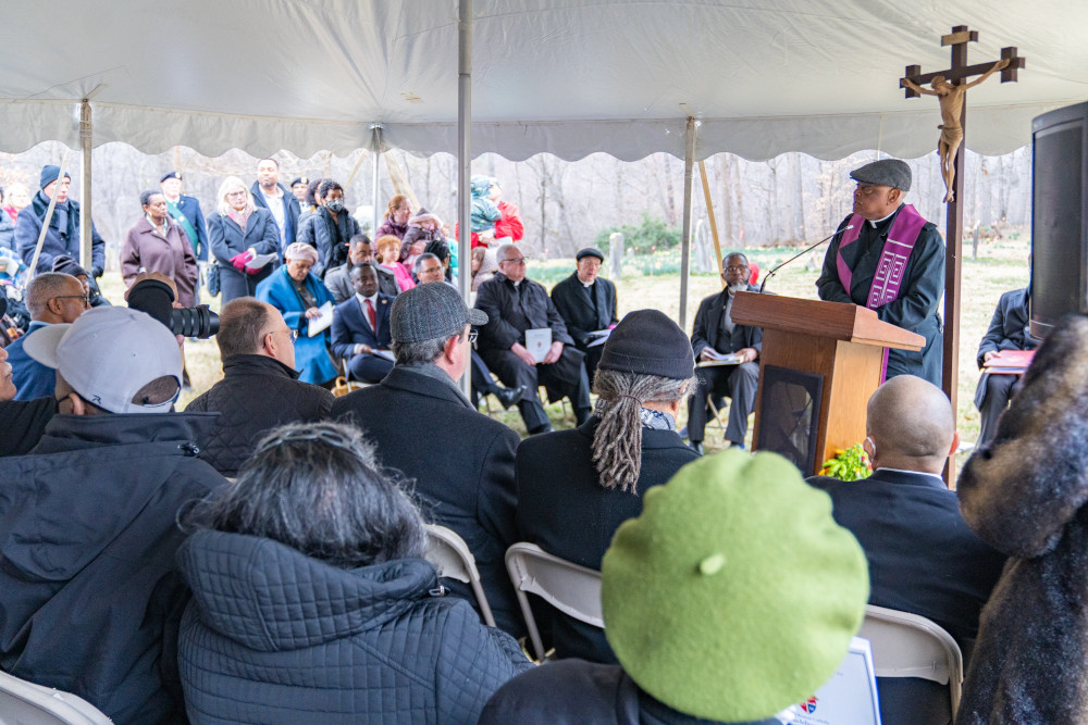 A Black man wearing a tweed cap, a black cassock, and a purple stole speaks to a large group of seated people under a tent. A crucifix is in the background