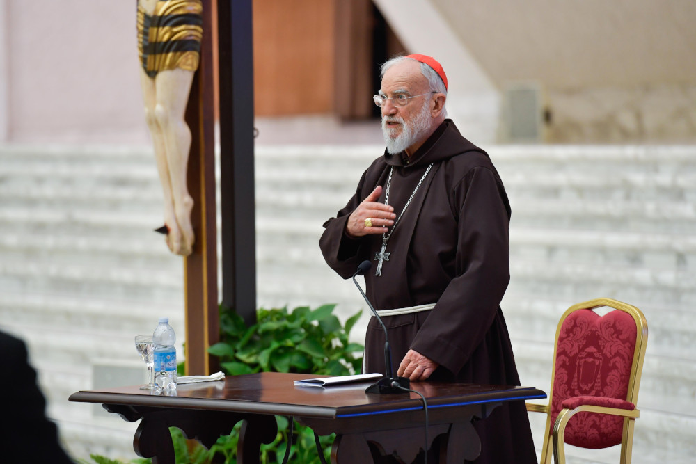 An older white man in a red zucchetto, pectoral cross and brown tunic with a chord speaks next to a crucifix