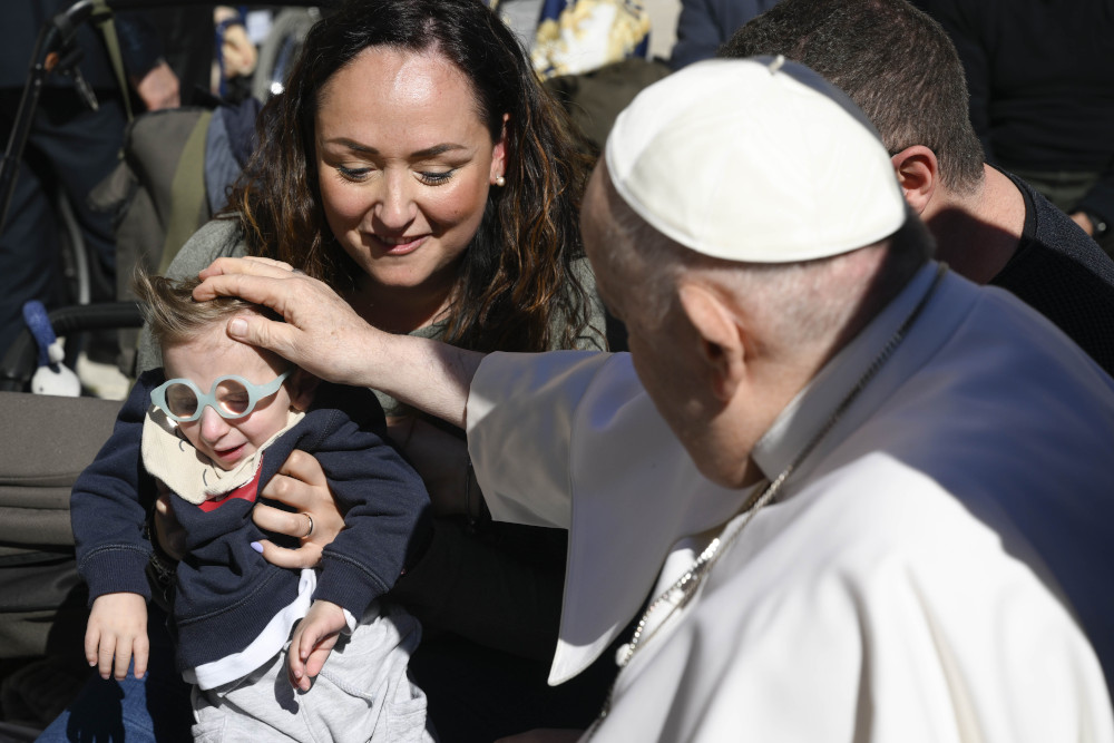 Pope Francis places his hand on a small child's head. The child has blue glasses and is held by a woman.