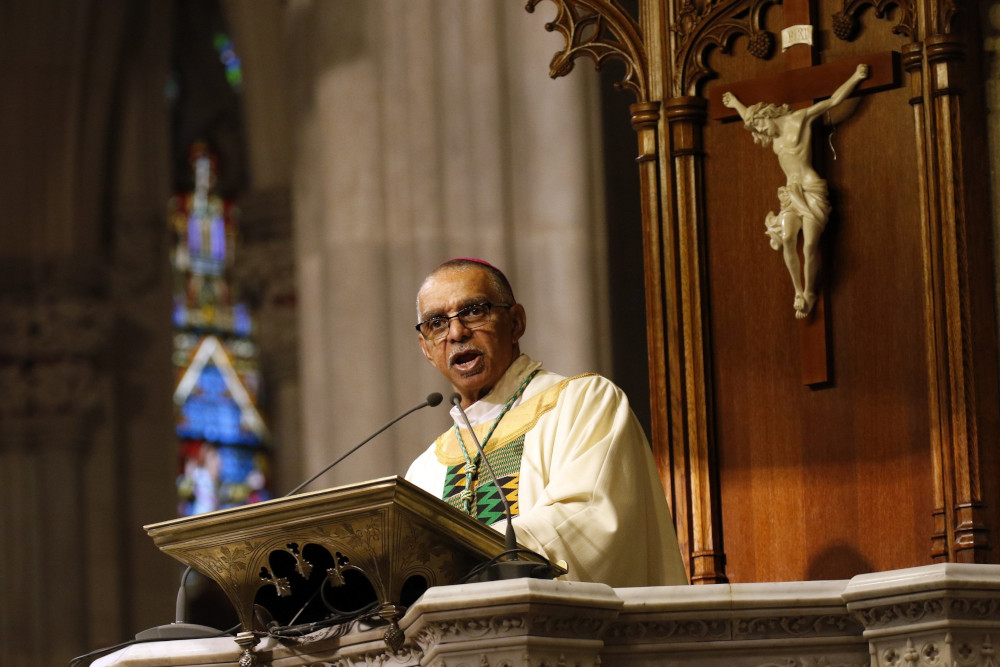 A light-skinned Black man speaks behind a lecturn and in front of a crucifix