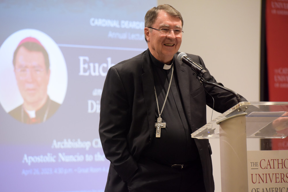 A white man in a black cassock wearing a silver pectoral cross speaks at a clear lecturn into a microphone