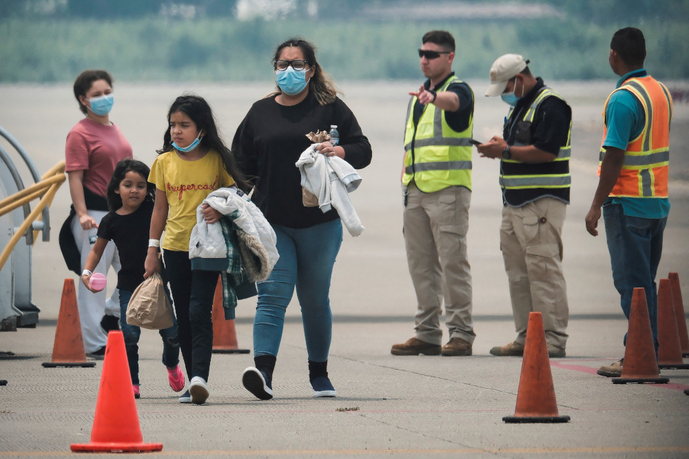 Brown people of various ages wear masks and stand on an airport tarmac next to men in safety vests