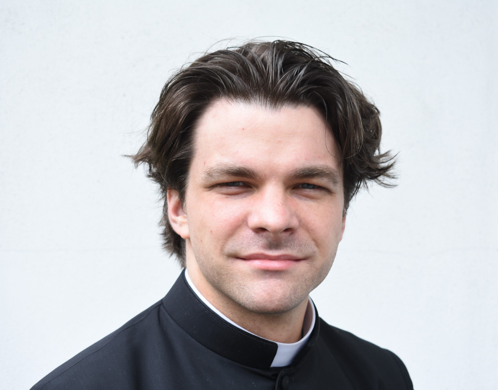 A white man wearing a clerical collar with styled, swoopy hair looks into the camera