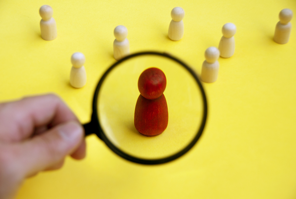 A hand with a magnifying glass singles out a red-painted wooden figurine among seven unpainted figurines (Dreamstime/Mauriceyom98)