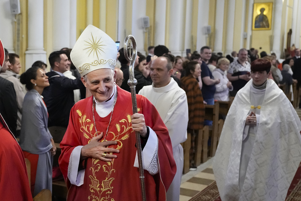 A man in a white mitre and red vestments walks down a church church aisle