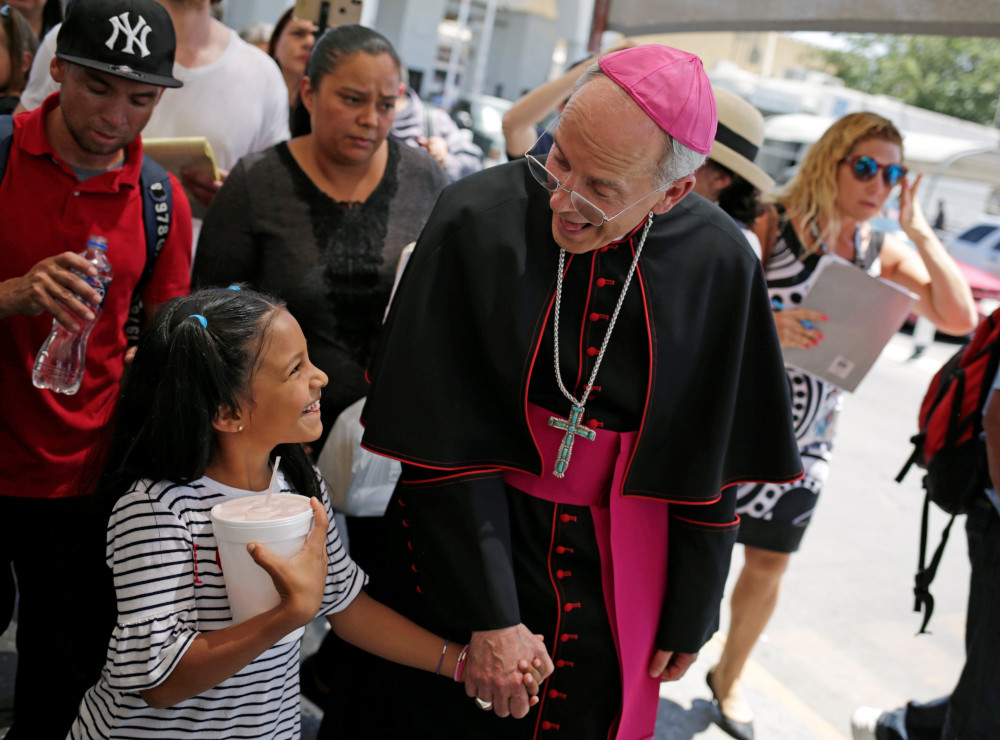 A white bishop wearing a house cassock looks down at a brown girl and holds hands with her as they smile at each other