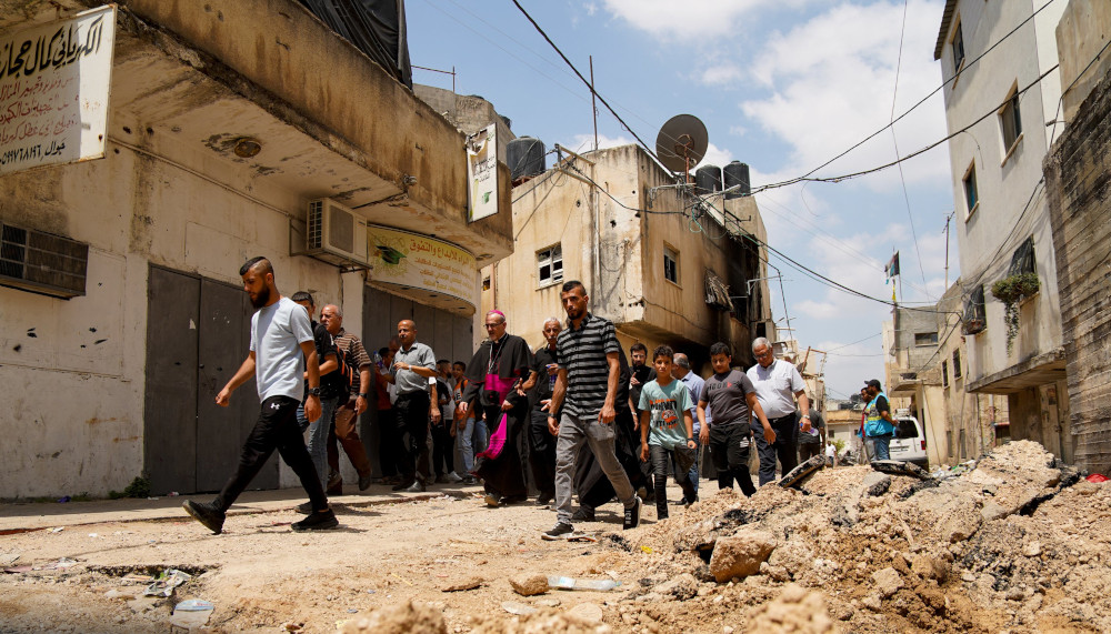 A man in a bishop's cassock walks with a large group of men between concrete buildings and rubble