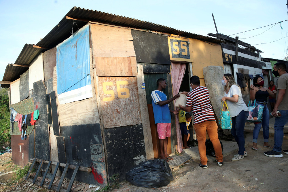 Masked people hold bags as they stand outside a house structure made of scrap wood and metal