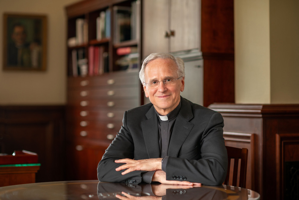 An older white man wears a clerical collar and black suit jacket in an office space