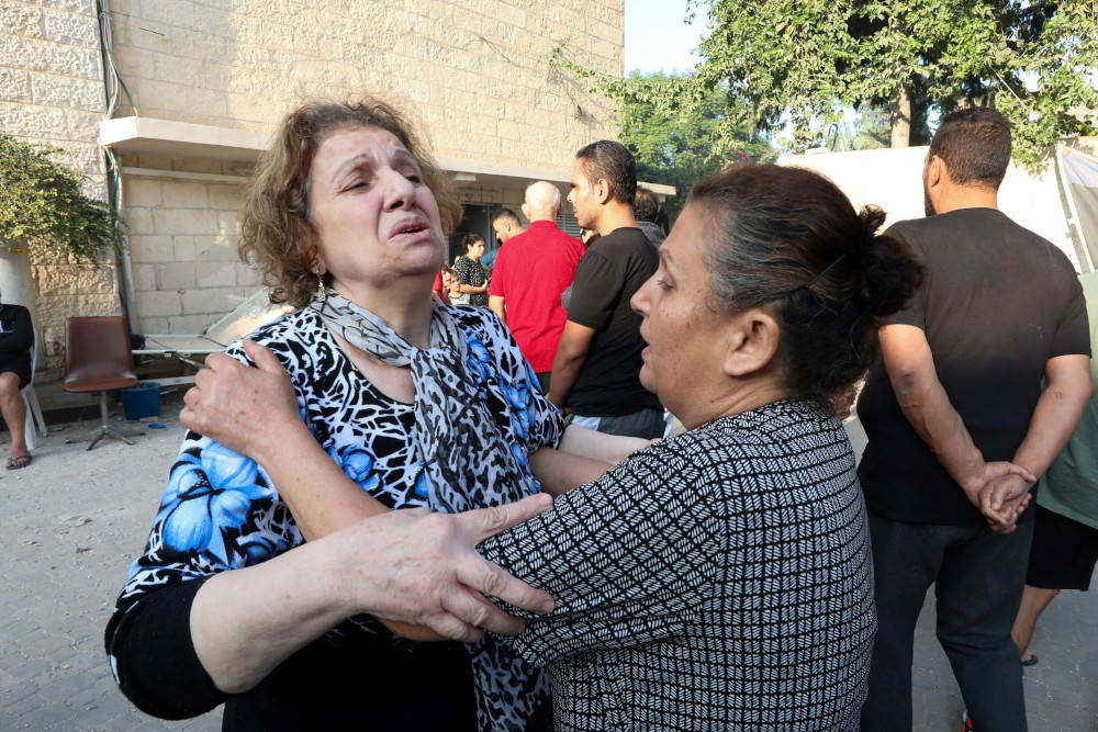 Two women, one with an extremely distressed facial expression, hold each others shoulders while standing outside among other peopel