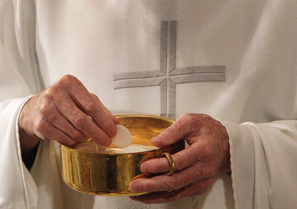 A white man's hands hold a pyx with Communion wafers. The man wears a white vestment with a cross on it.