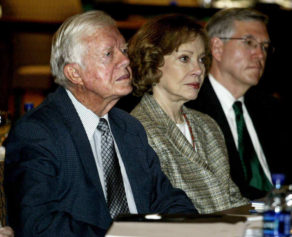 A brown-haired older white woman sits between two older white men. All wear formal clothes.