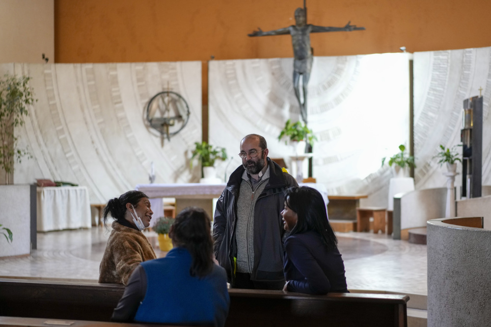 Three women sit in pews in a church while a casually dressed priest speaks with them