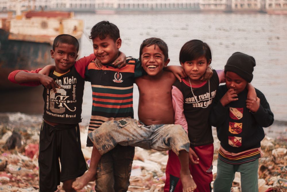 A group of children stands in a line, arms across each other's shoulders. A body of water is behind them. 