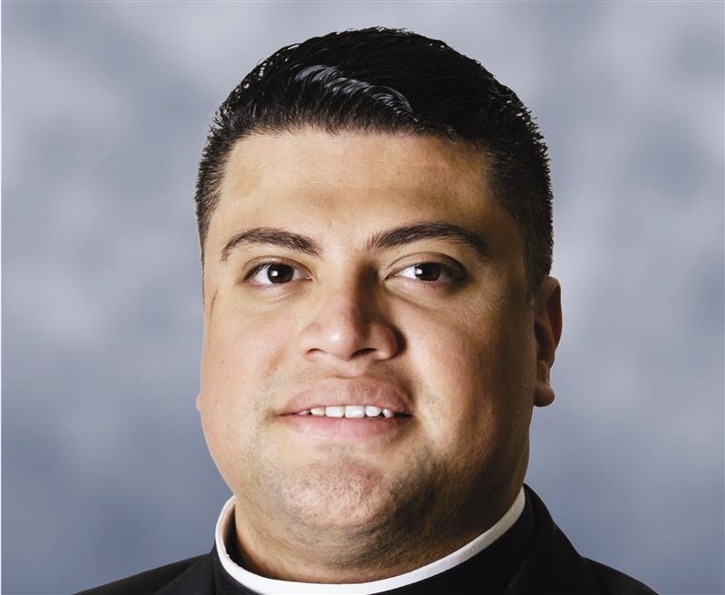 A Latino man with short hair and a clerical collars looks into the camera with a slight smile