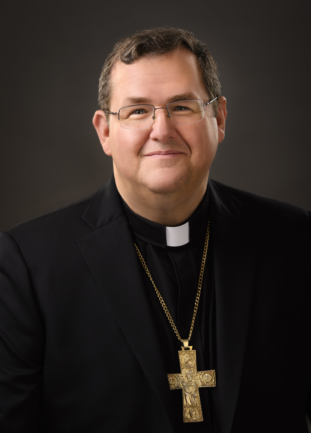 A white man with brown hair and glasses smiles at the camera. He wears a clerical collar and a pectoral cross.