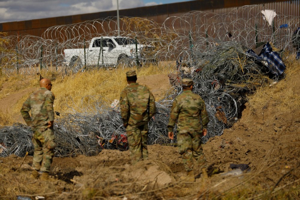 People in camo uniforms stand next to twisted razor wire. Clothing is caught in different parts of the razor wire.