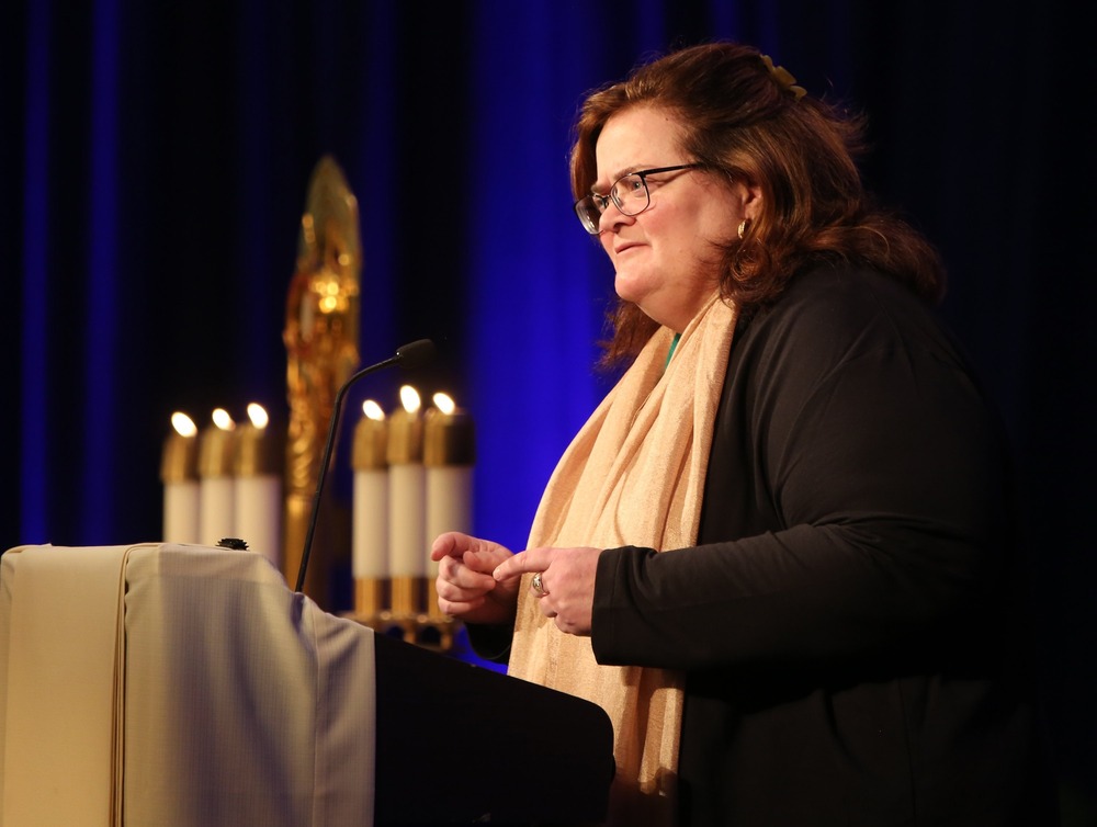 Teresa Pitt Green stands at lectern, speaking with candles in background.