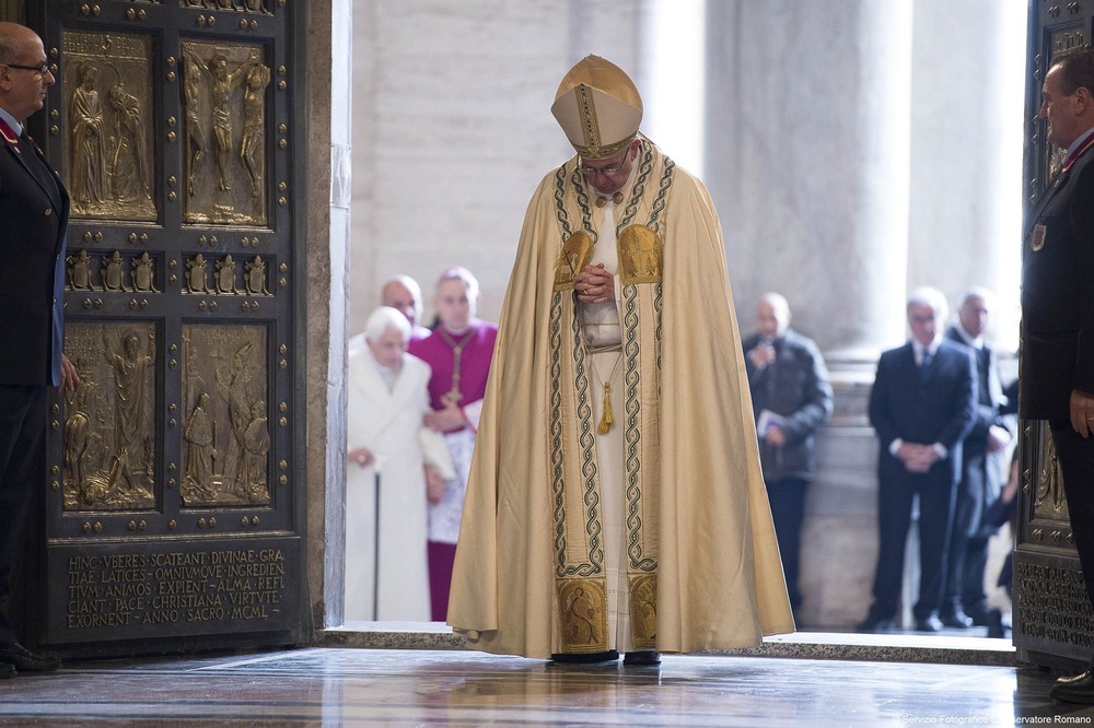 Pope Francis, vested, walks with head bowed through Holy Doors of St. Peters; in background is Pope Emeritus Benedict XVI, wearing plain white coat, supported by cane and bishop. 