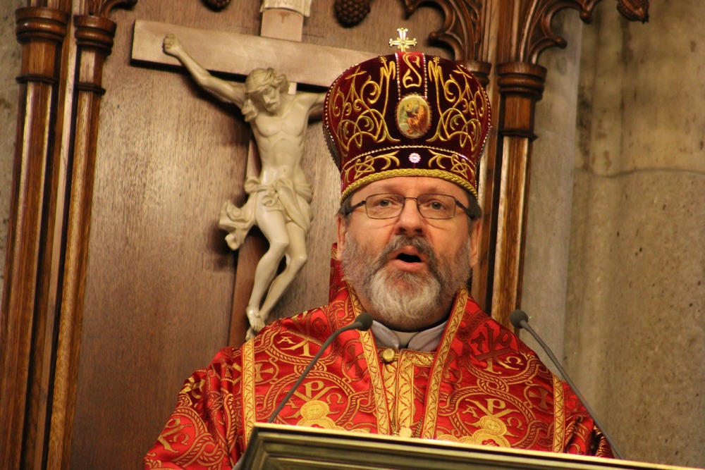 Bishop, vested in eastern style, delivers homily from lectern, crucifix in background. 