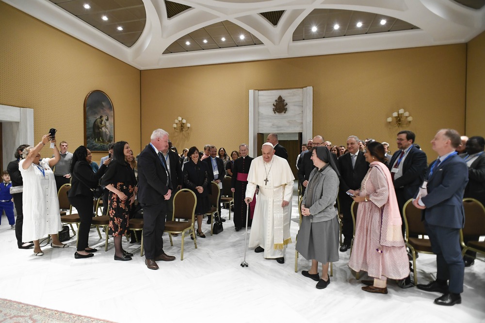 Pope Francis stands among conference attendees in large hall. 