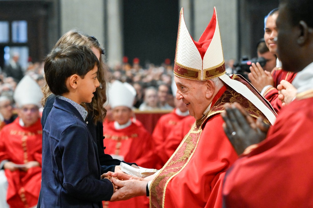 Pope Francis, seated and vested in red, bends and smiles tenderly to two small children. 