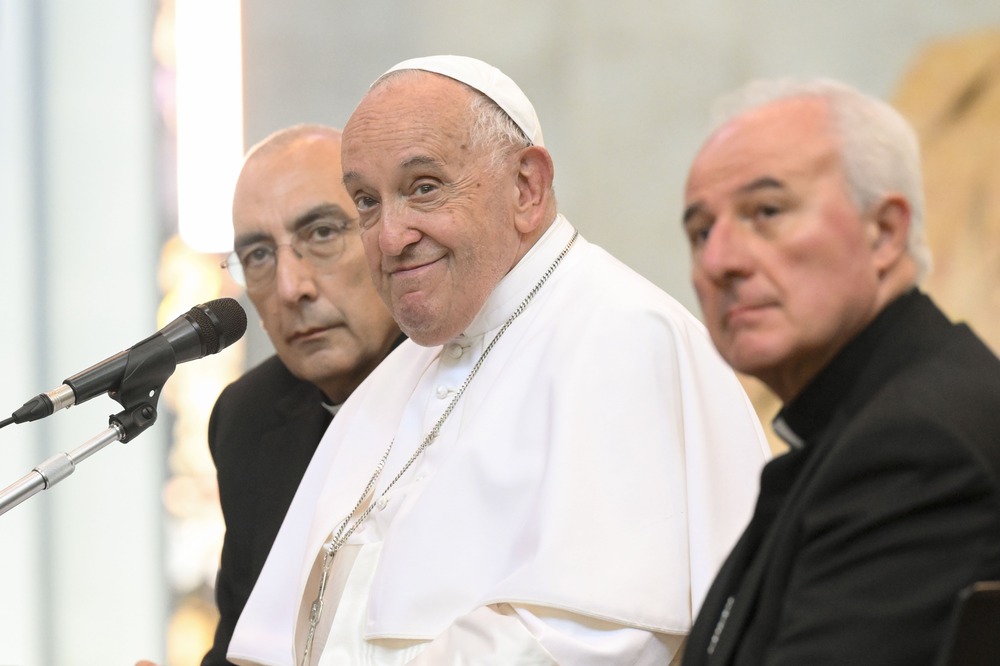 Pope Francis, seated, smiles as he listens. 