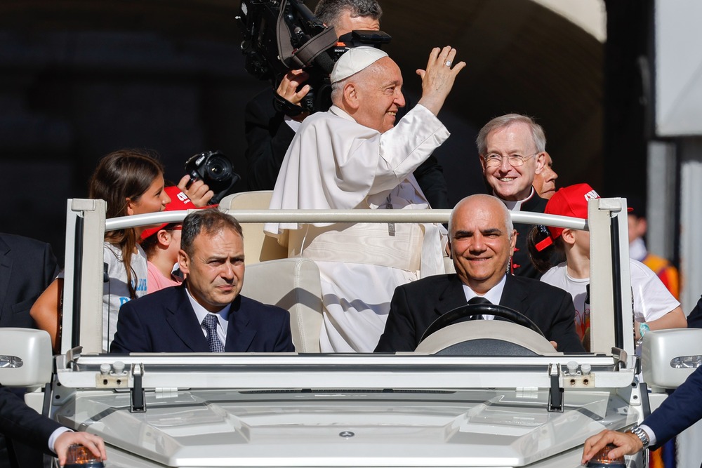 Pope Francis, standing in popemobile and framed by driver and attendants, waves to crowds