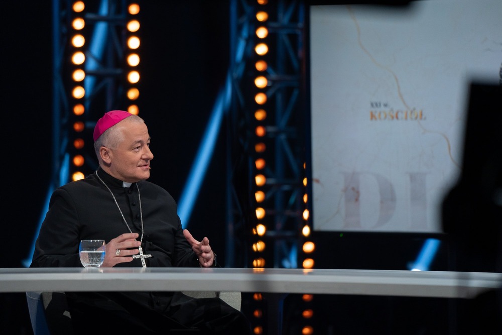 Bishop, in black cassock and purple zucchetto, talks on television. 