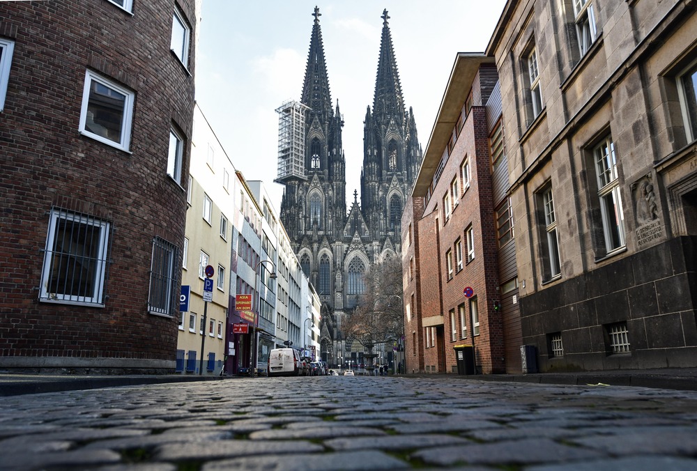 Cathedral of Cologne, one spire obscured by scaffolding, looms over cobblestone street.