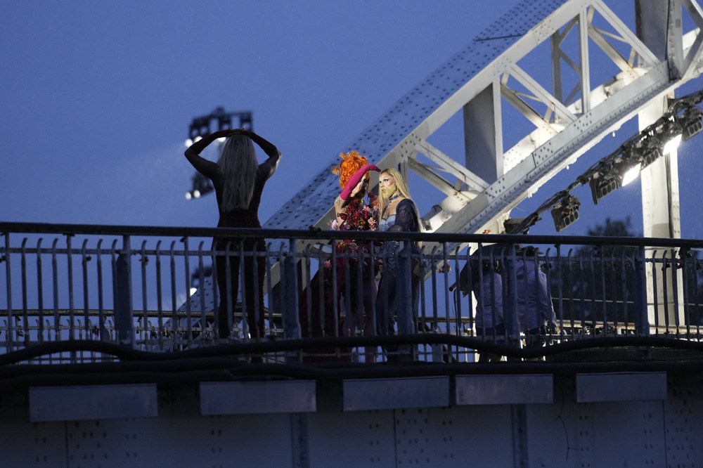 Two queens stand on bridge, dimly lit from behind, silhouetted camera operator visible in foreground. 