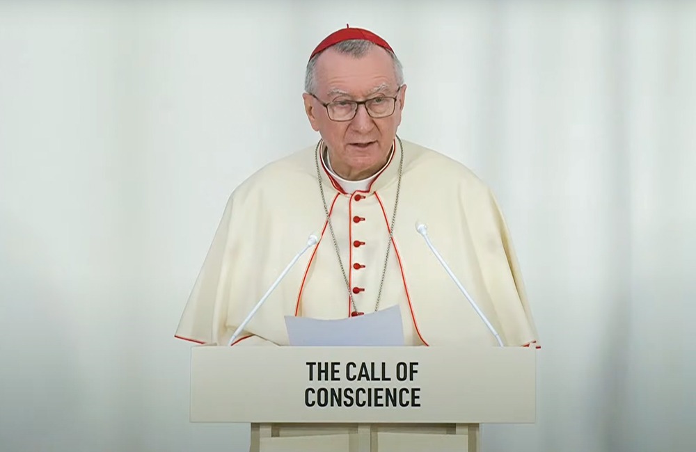 Parolin wearing white cassock piped with Cardinal's red and red zucchetto stands at podium reading remarks. 