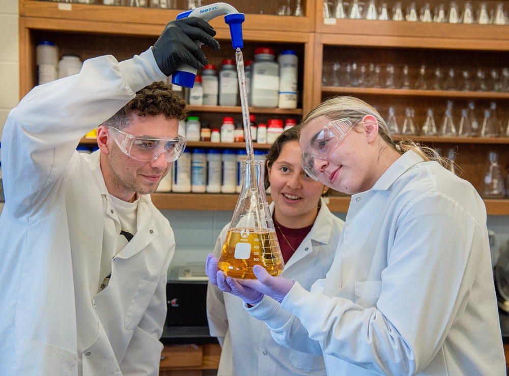 Three students wear white lab coats, and observe as one student uses a dropper to insert fluid into Erlenmeyer flask. 