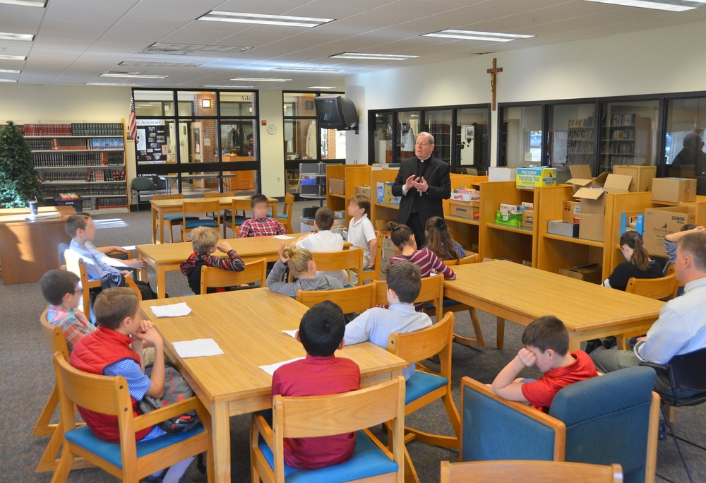 Children sit at table in large room, while priest stands talking. 