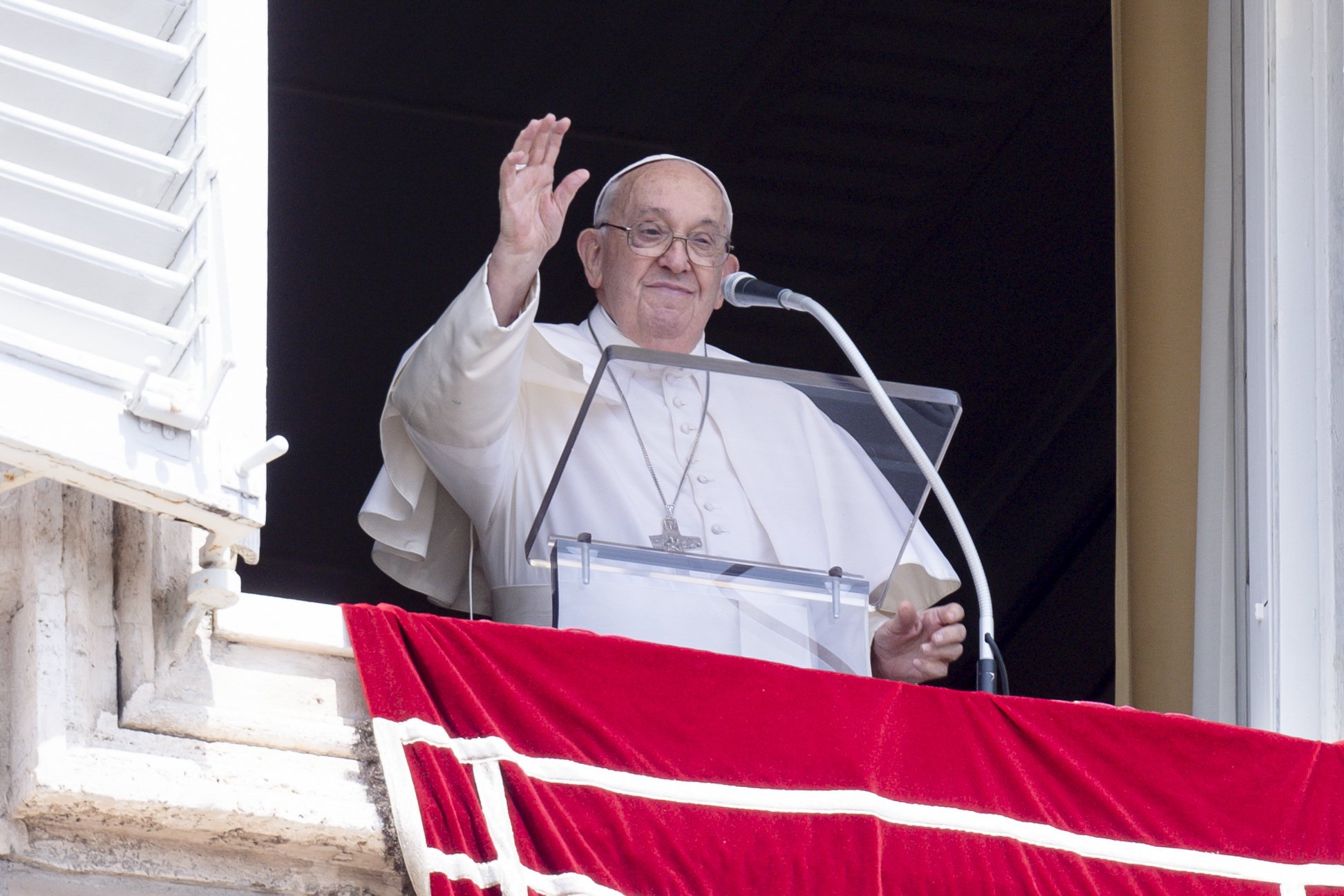 Pope raises hand in greeting, standing at lectern in window of Apostolic Palace. 