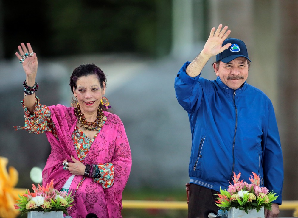 The Presidential couple stand waving and smiling in open air. 