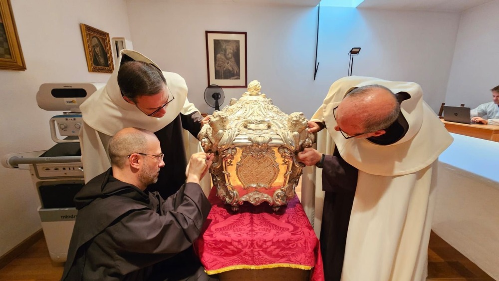 Three men religion in habits bend down to look at elaborate silver coffin.