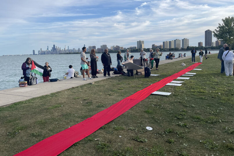 Daylight at lakeshore with Chicago skyline in background; long red ribbon stretches across entire frame. 