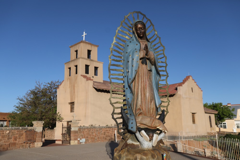 Statue of Our Lady of Guadalupe shown in front of large pueblo Church.