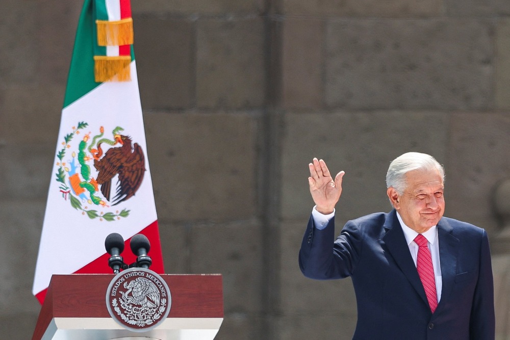 Obrador raises hand in greeting, standing on a dais; behind him is a podium and a Mexican flag. 