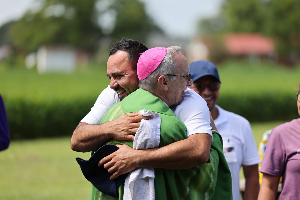 The bishop, vested in green, hugs a smiling man; in the background is a field of tall green stalks, possible corn. 