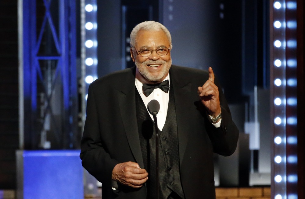 Jones, wearing formal wear, speaks from stage; hand raised in gesture, beaming a large smile.