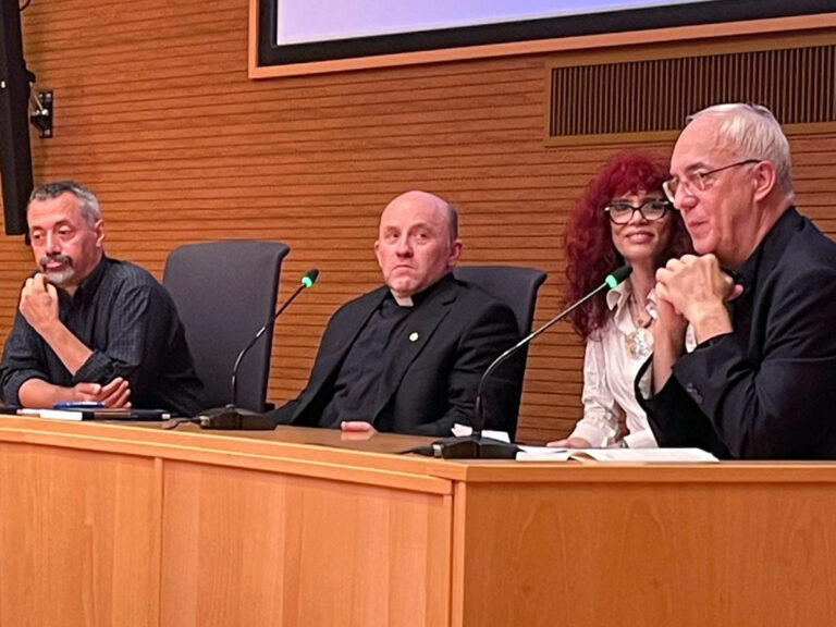 The four sit at a wooden desk with microphones. 